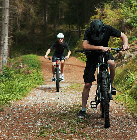 Zwei E-Bike-Fahrer auf einem Waldweg, der vordere hält an und blickt nach unten