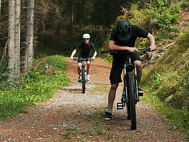 Zwei E-Bike-Fahrer auf einem Waldweg, der vordere hält an und blickt nach unten